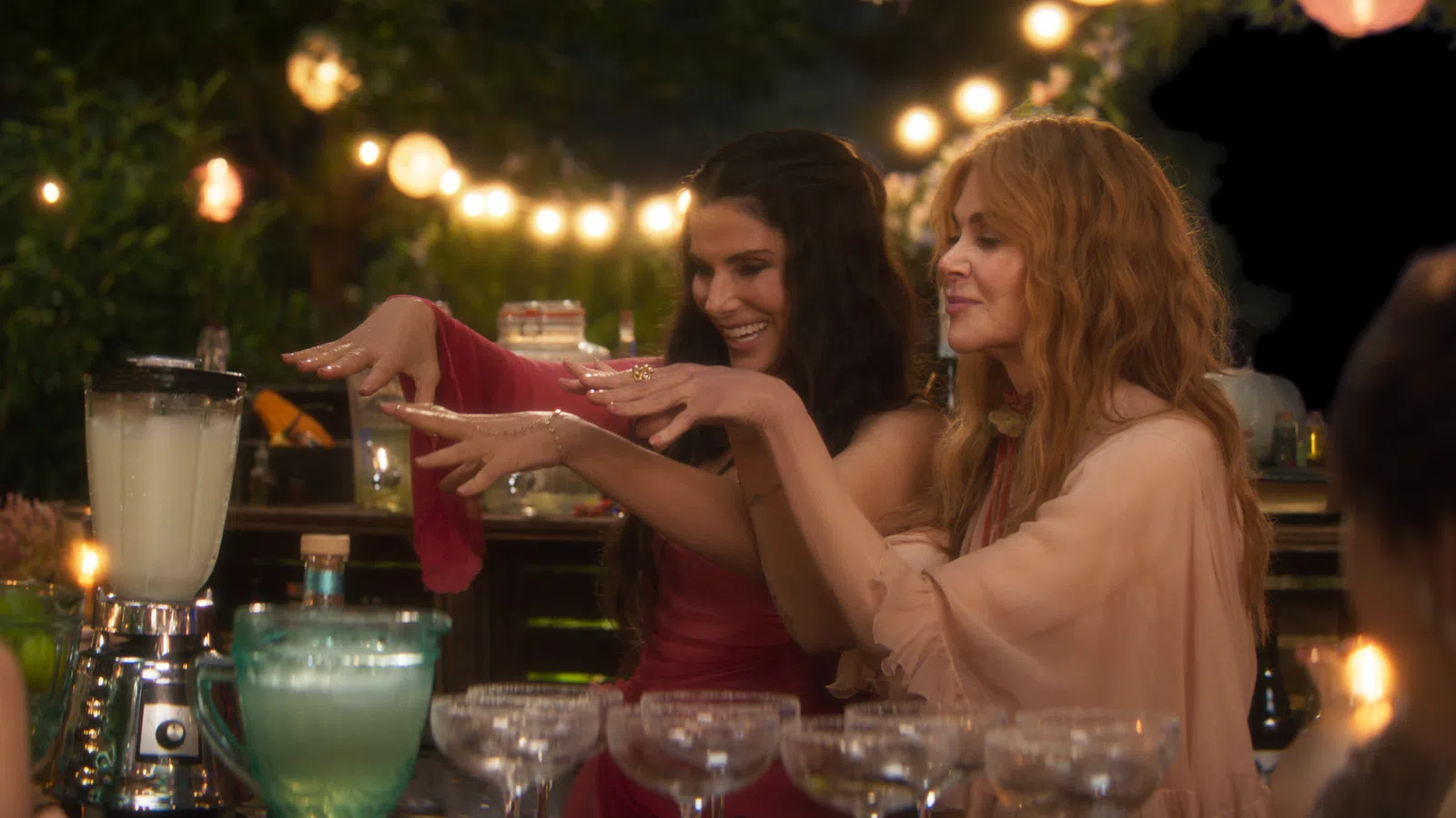 Two women at a festive outdoor bar preparing drinks with string lights and glassware in the background.