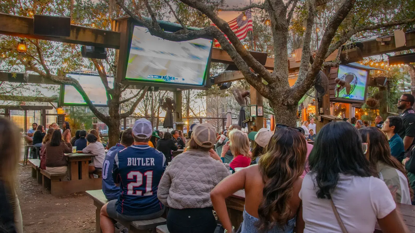 Outdoor sports bar with crowd watching a football game on multiple large screens under trees and string lights.