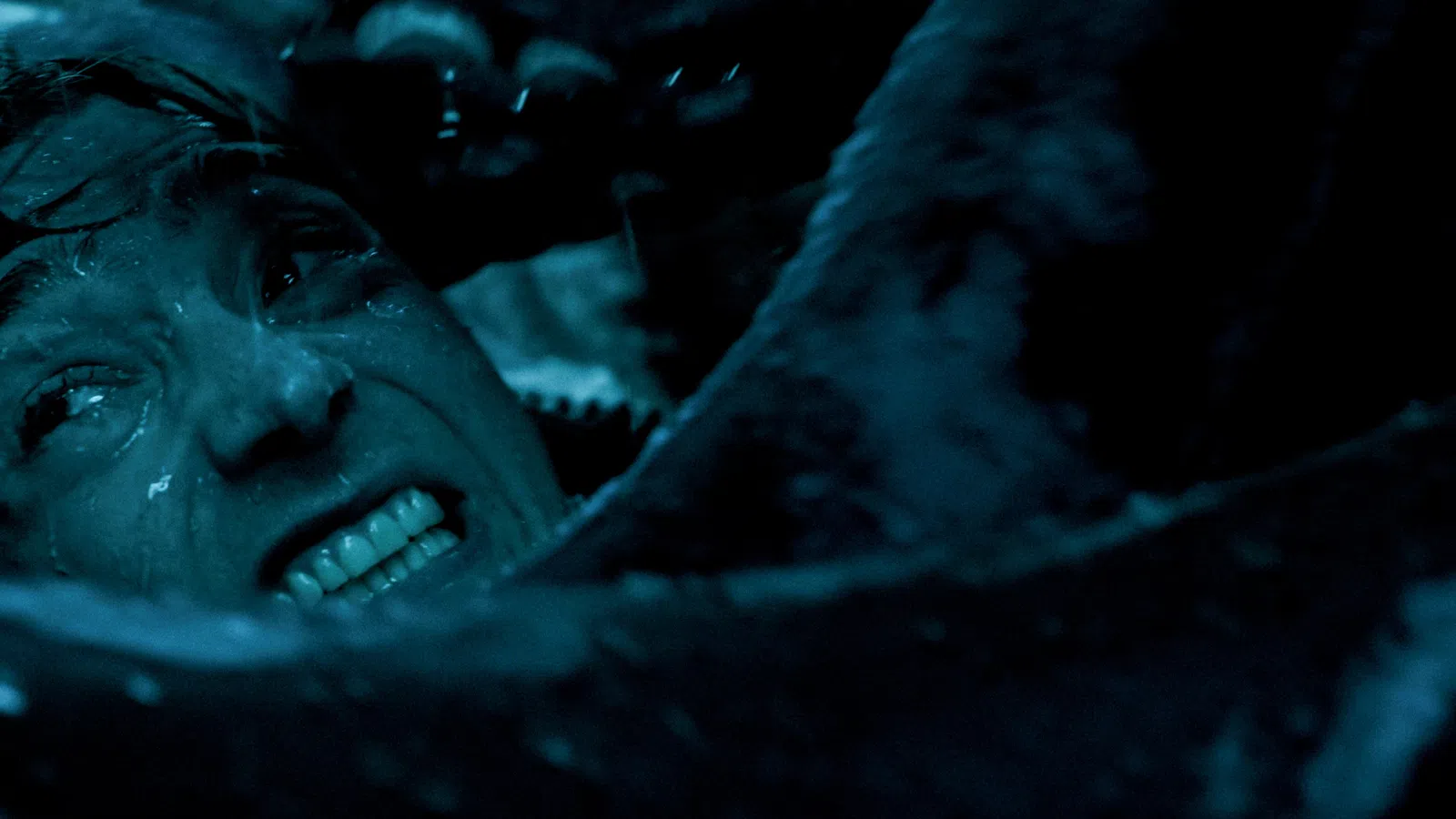 Close-up of a man's tense face underwater surrounded by tentacles in dark blue lighting.