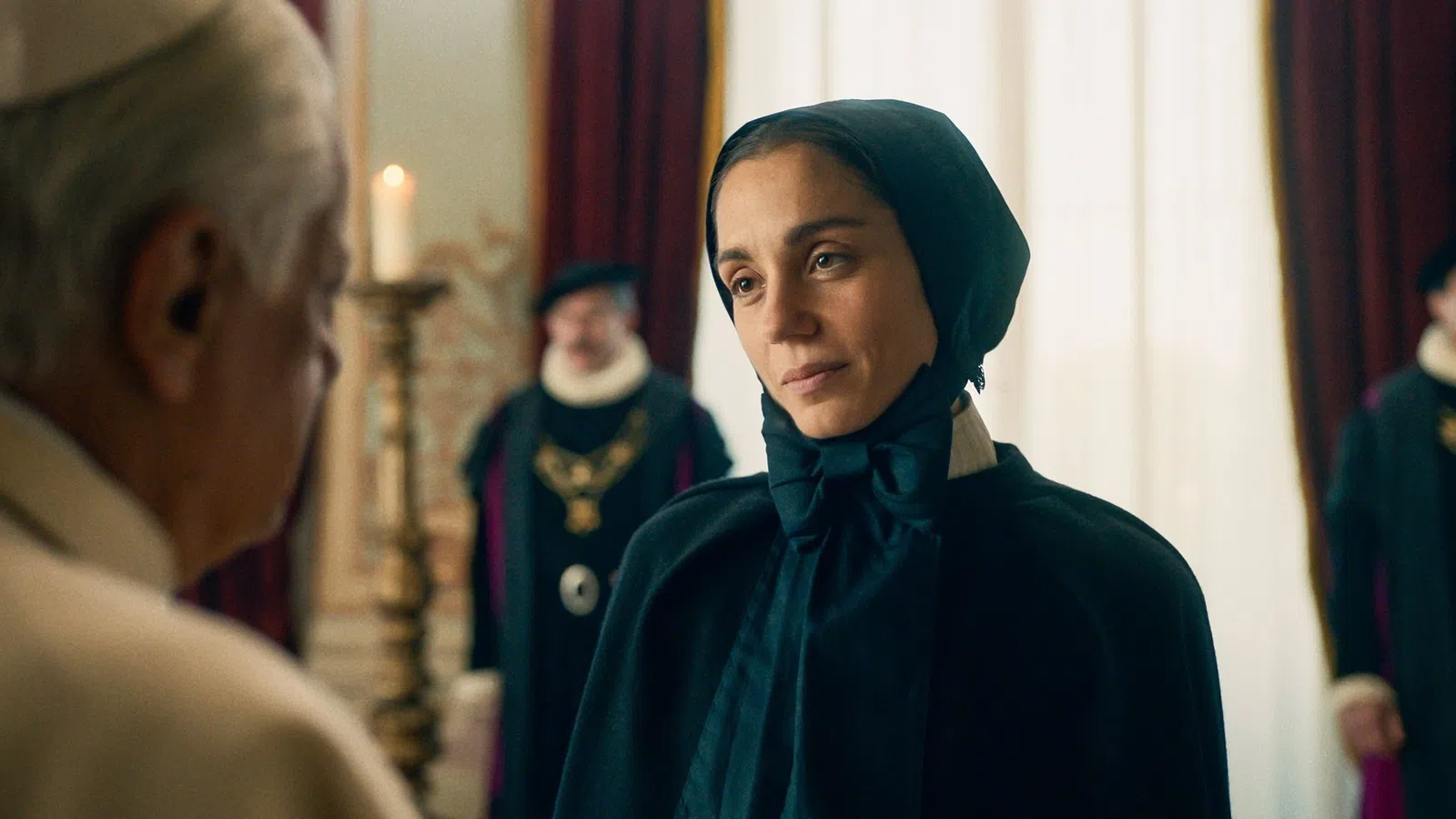 Woman in black medieval attire looking thoughtfully at an older man in a royal setting with candle and attendants.