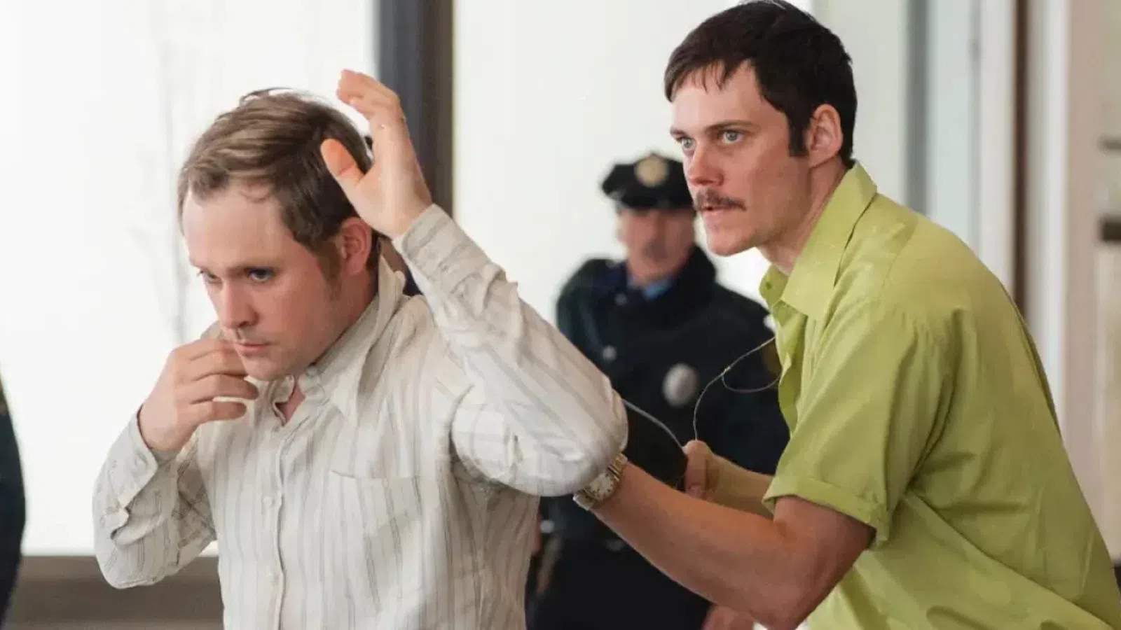 Man in striped shirt being handcuffed by another man in green shirt with police officers in background