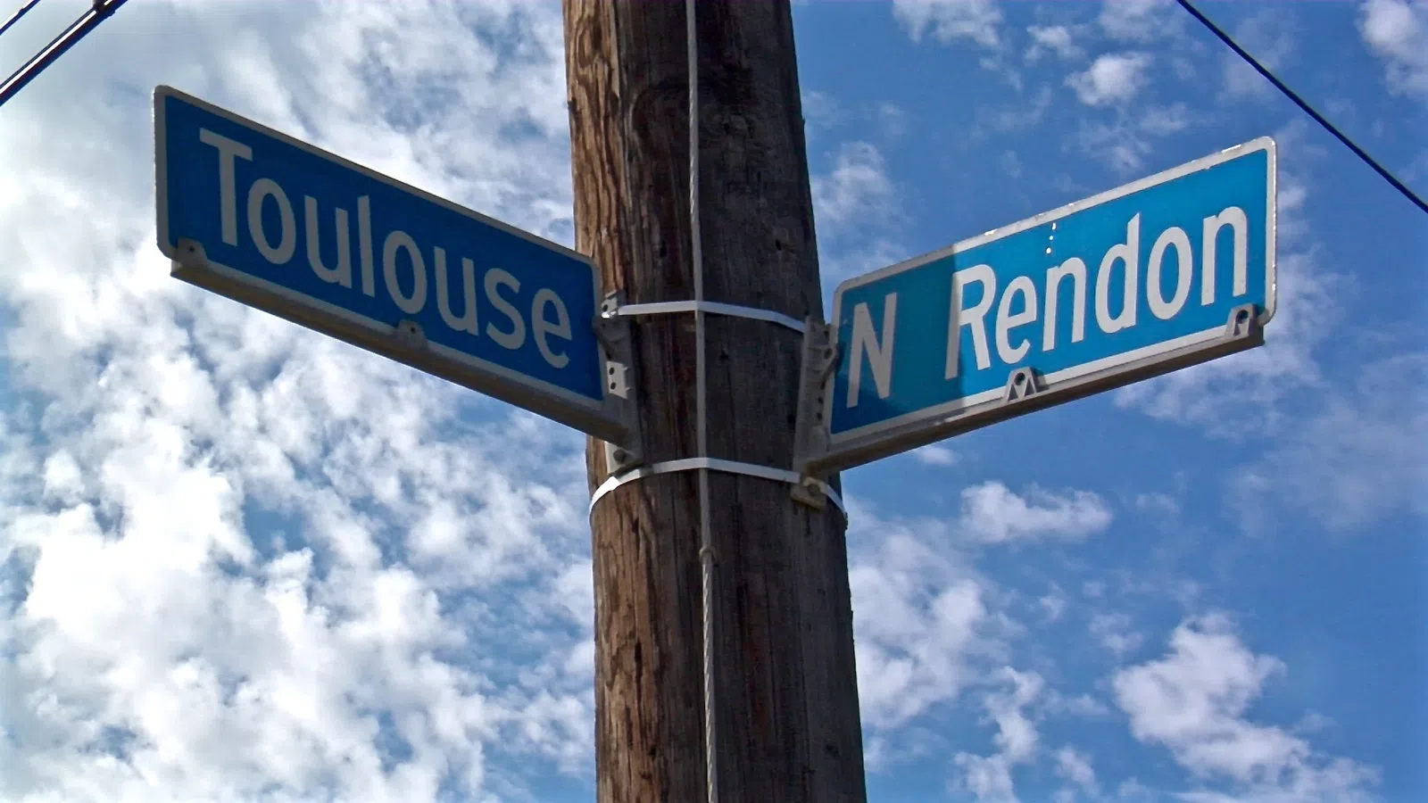 Street signs for Toulouse and N Rendon mounted on a wooden pole against a partly cloudy blue sky.