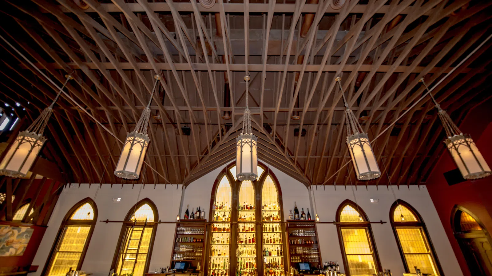 Upscale bar with gothic arched windows, illuminated shelves of liquor, and wooden ceiling beams with hanging lanterns.