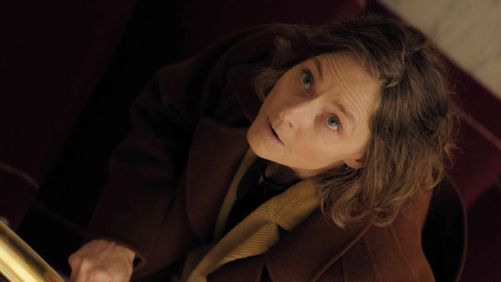 Woman in brown coat looking upward with a concerned expression in a dimly lit setting.