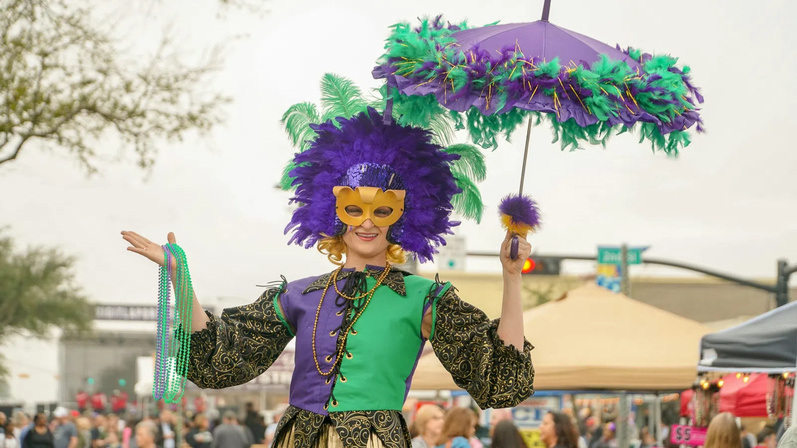 Person wearing a colorful Mardi Gras costume with purple and green feathers, mask, beads, and decorated umbrella.
