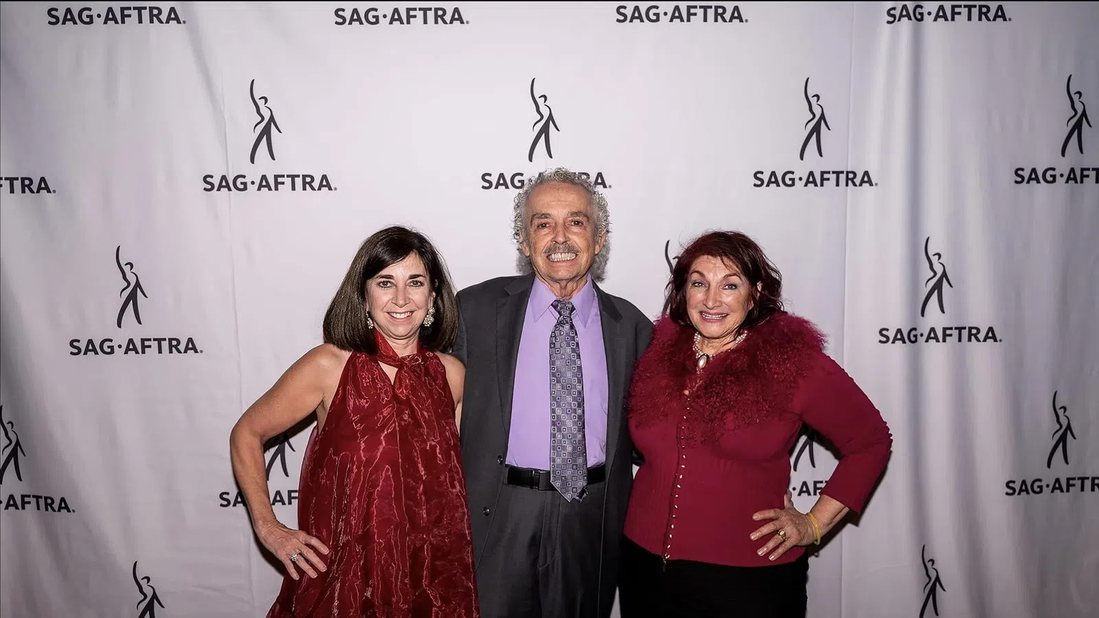 Three adults posing at SAG-AFTRA event with branded backdrop, two women in red outfits and one man in suit.