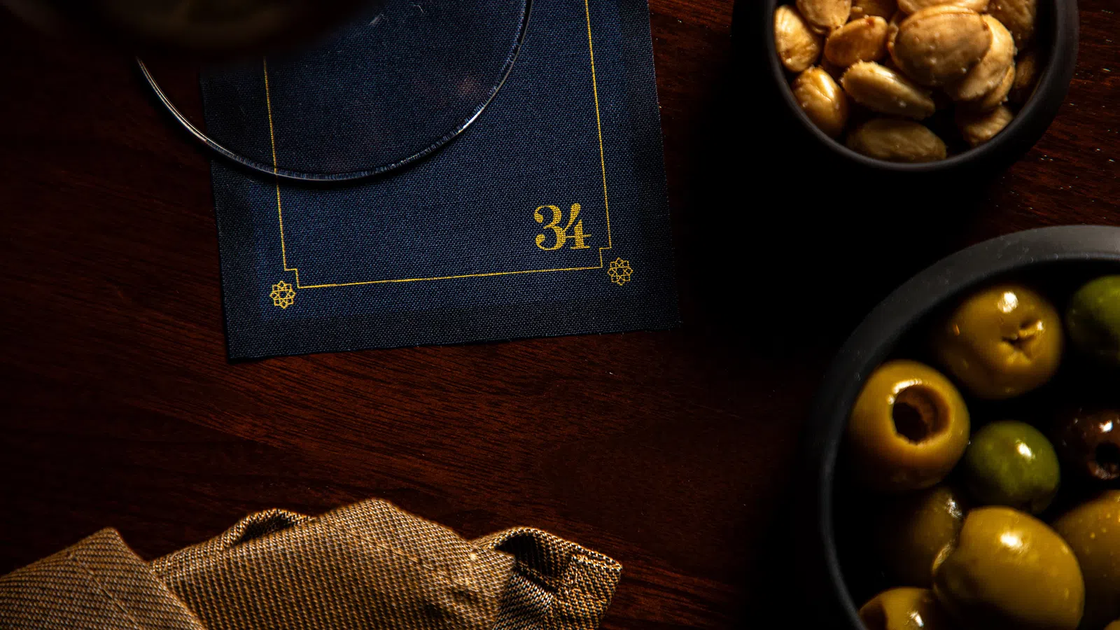 Dark wood table with bowls of green olives and roasted nuts beside a navy blue napkin numbered 34