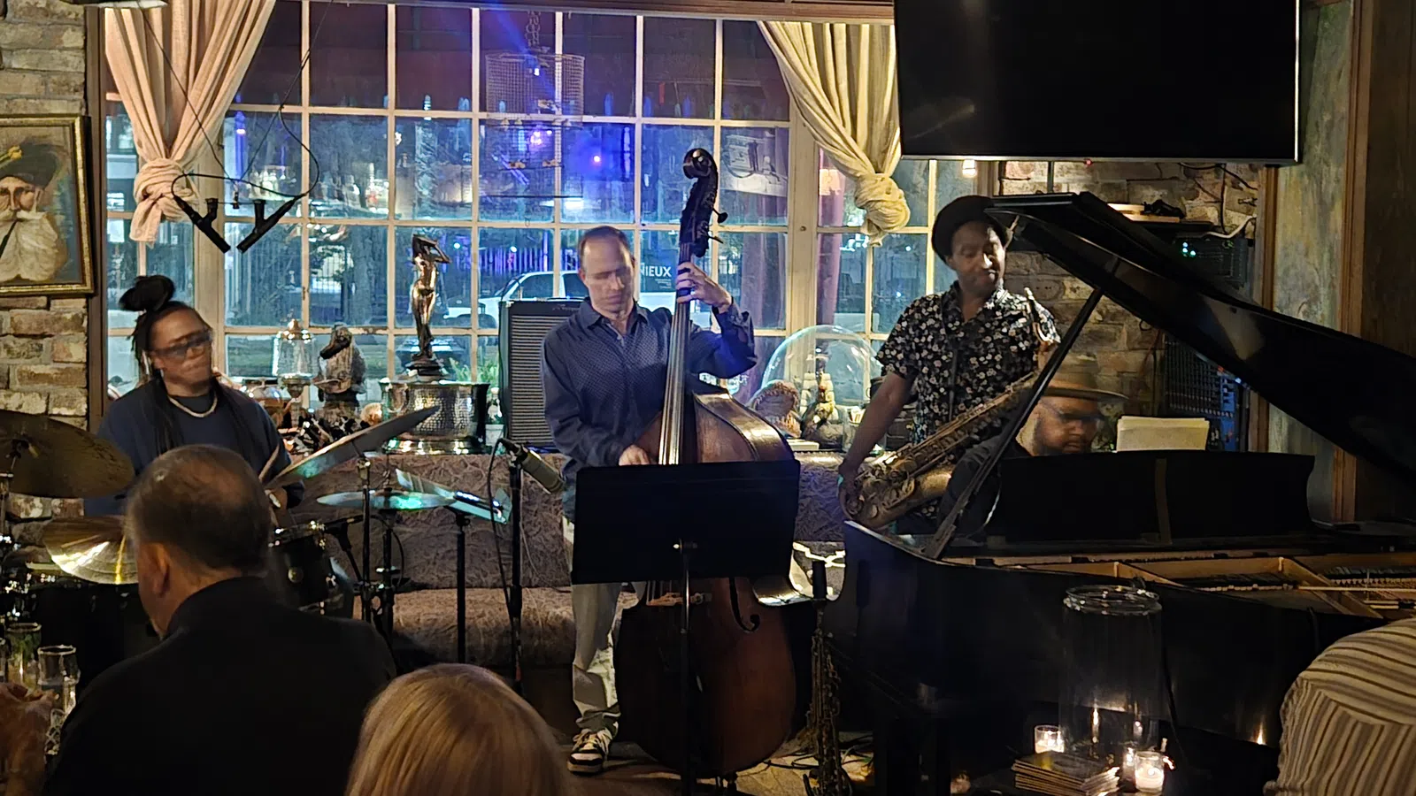 Live jazz band performing with drums, upright bass, saxophone, and piano in cozy pub setting with audience