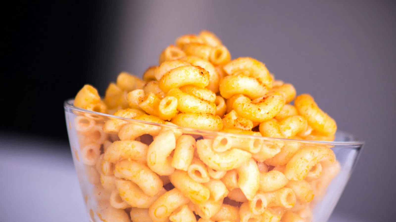 Clear glass bowl filled with curly, orange seasoned cheese snacks on a white surface with blurred background