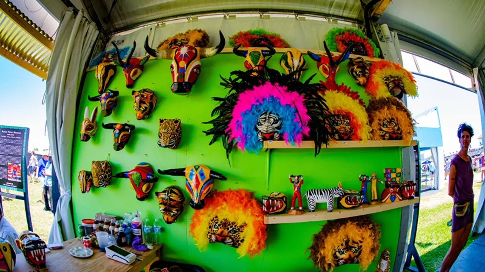 Colorful animal masks and crafts displayed on a bright green wall at an outdoor market stall under a tent.