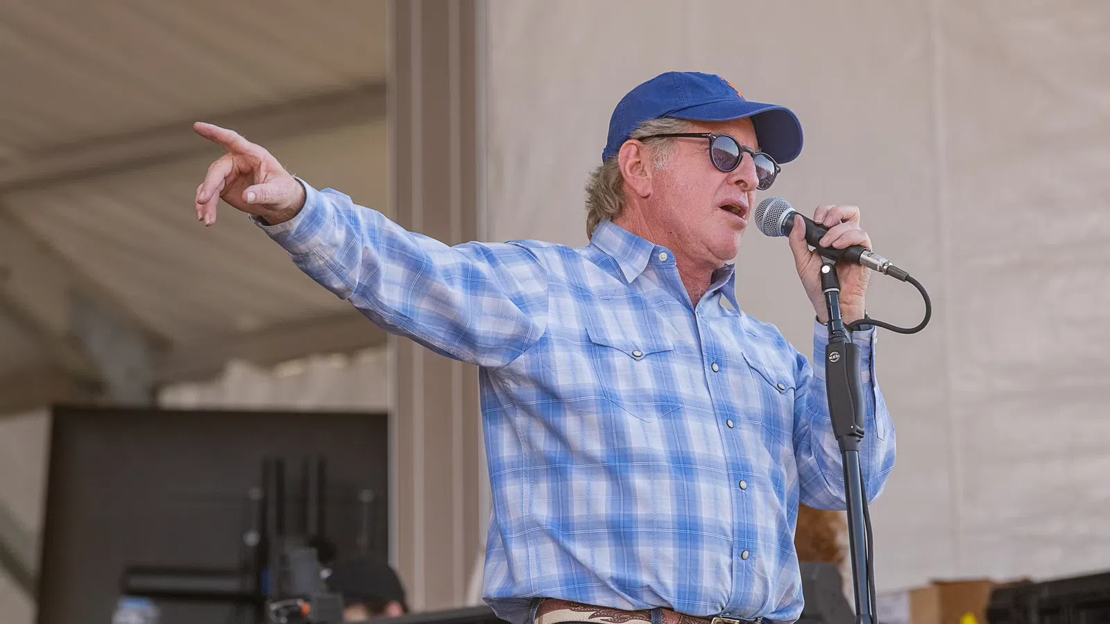 A man singing passionately on stage, wearing sunglasses and a blue shirt, with an audience in the background.