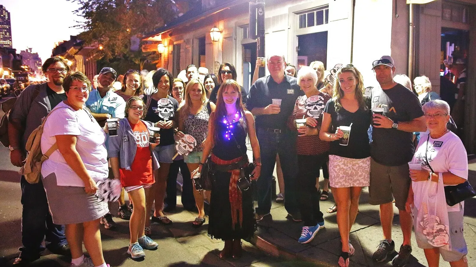 Group of people gathered on a lit street at night holding drinks and smiling, casual and festive atmosphere.