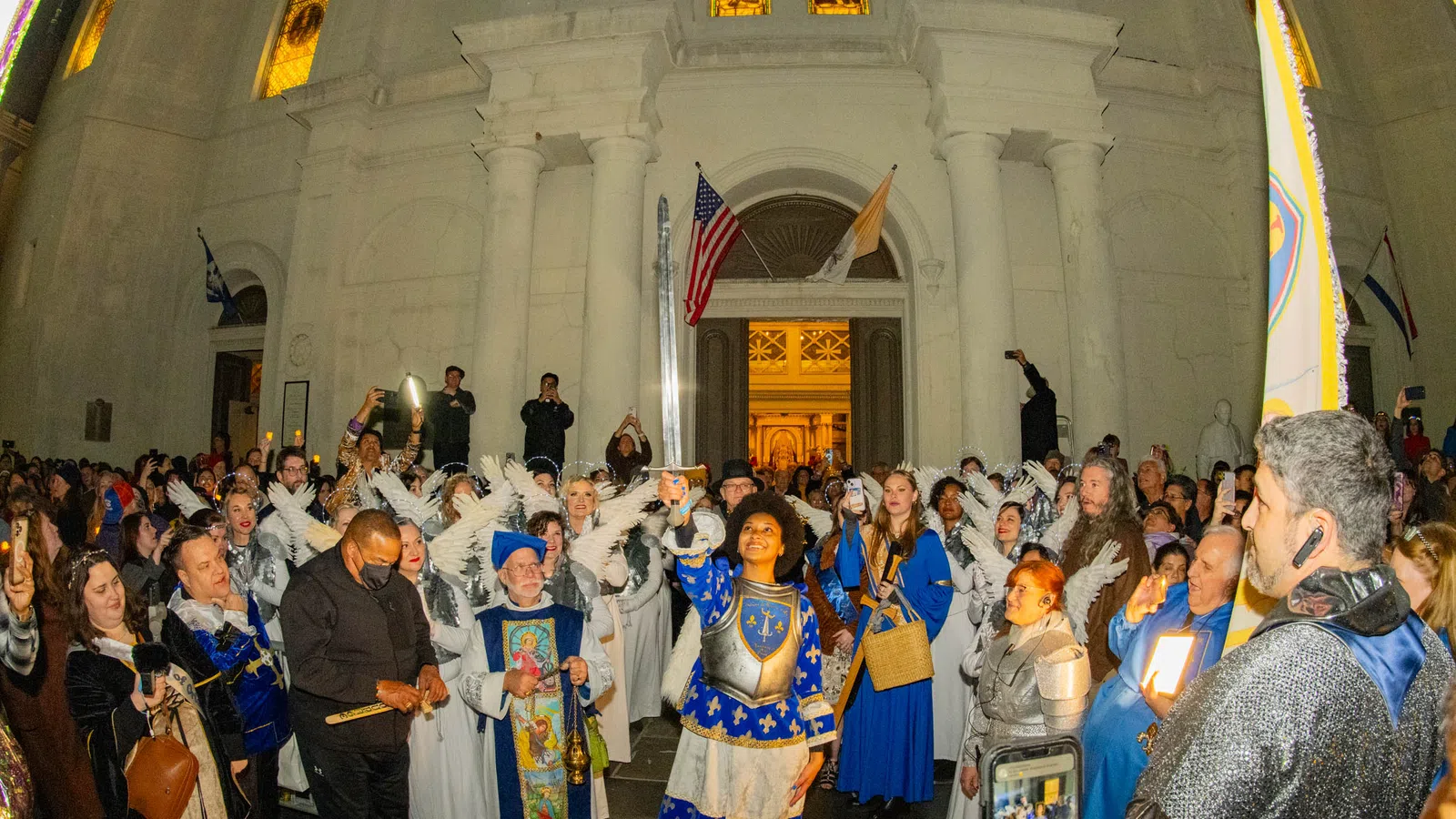 Nighttime religious procession with people in medieval costumes and angel wings outside a historic white church.