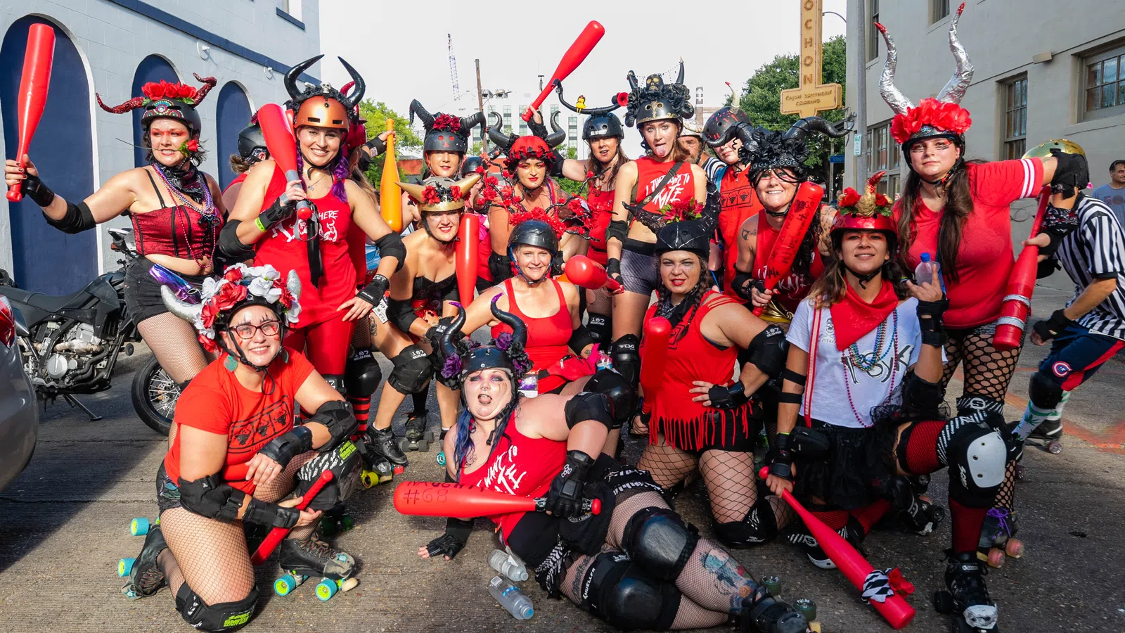 Group of roller derby players in red outfits and helmets posing with red bats on city street during daytime