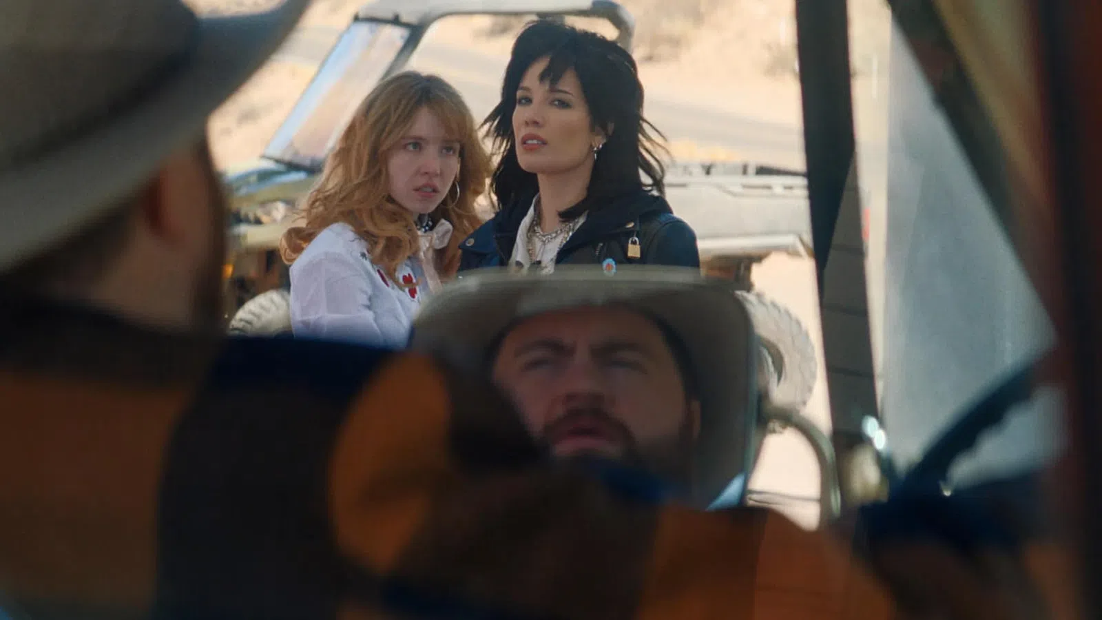 Two women stand outside a vehicle in a desert setting while a man in a hat is seated inside the vehicle.