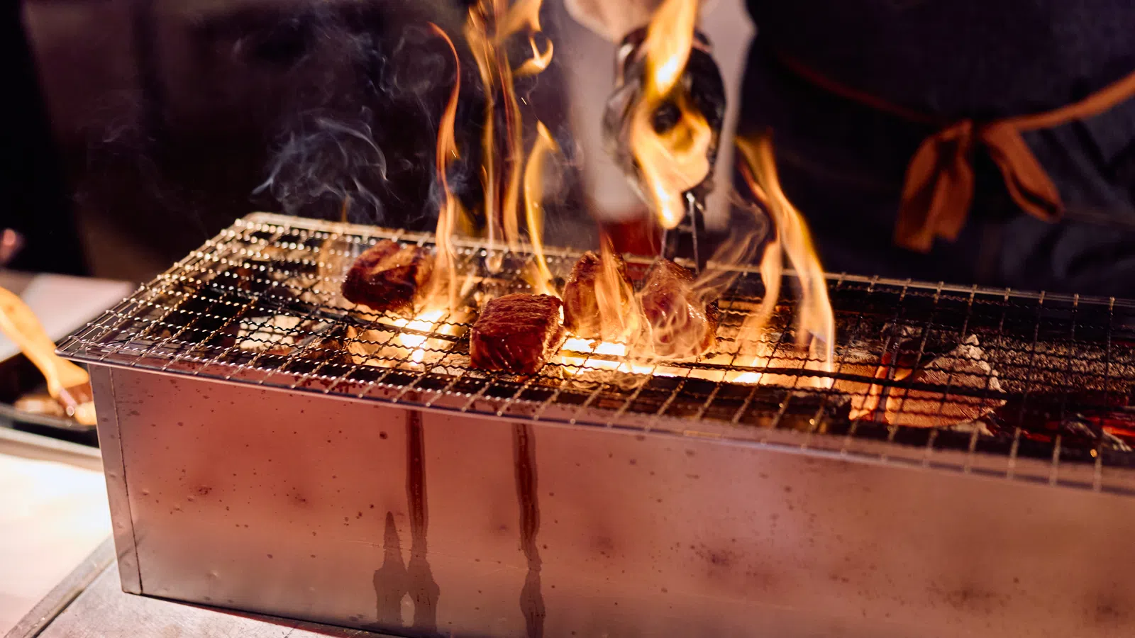 Flames flare up as meat pieces sizzle on a metal charcoal grill with a person grilling using tongs.