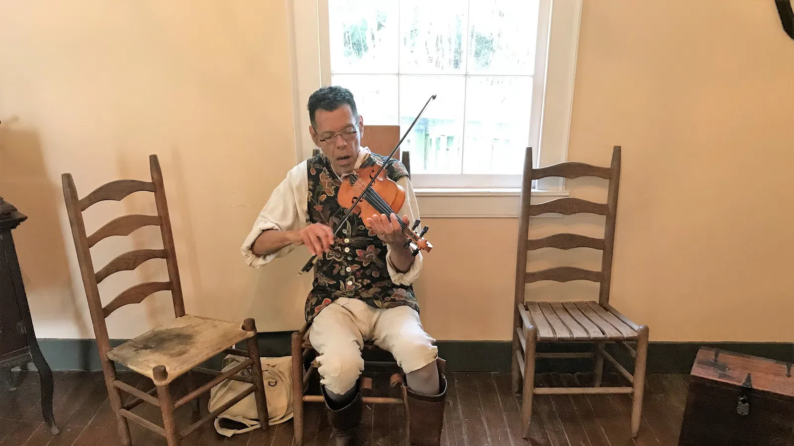 Man in historical costume playing violin while seated between wooden chairs on hardwood floor