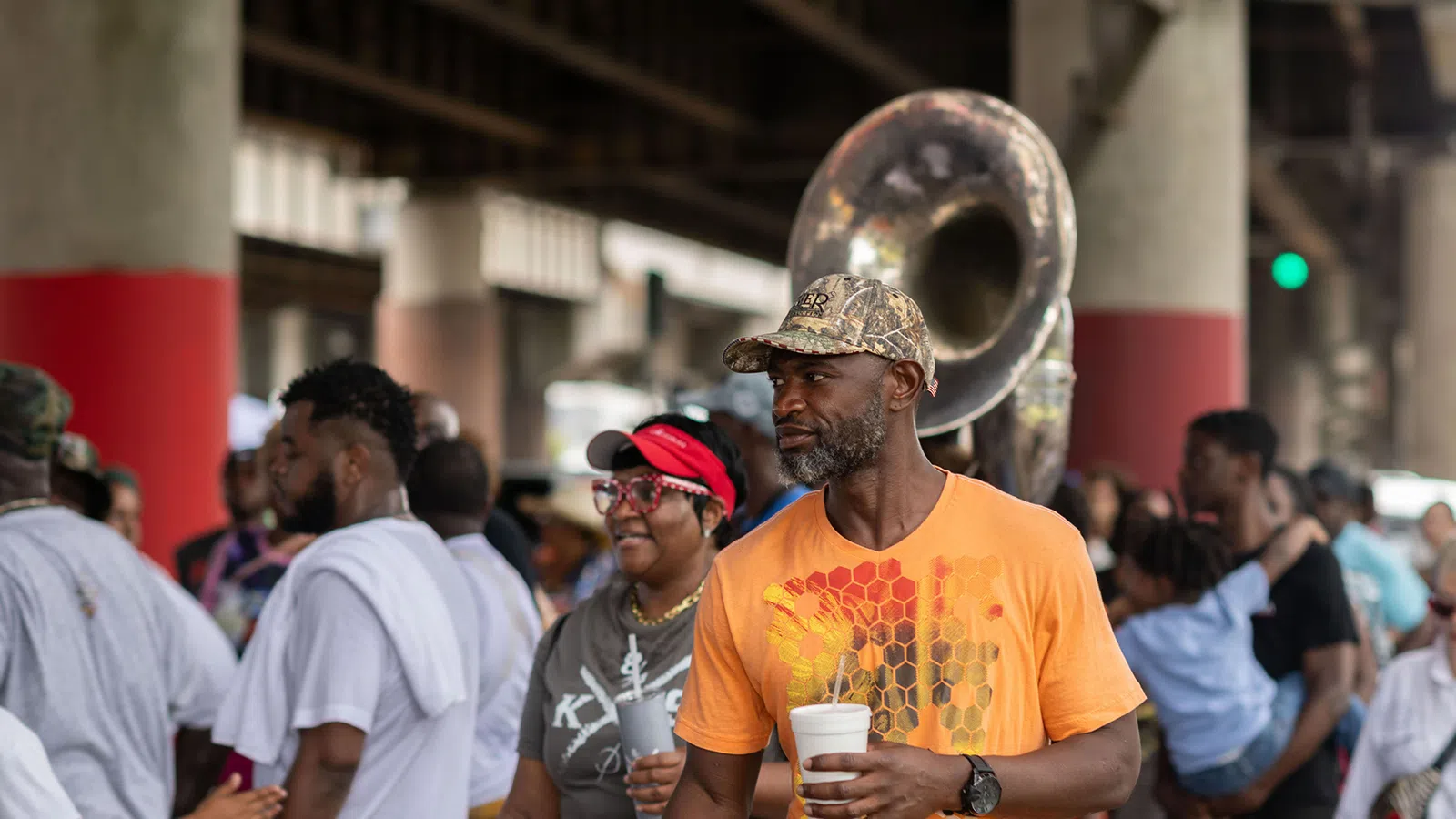 Man in orange shirt holding a cup with people and a sousaphone band under an overpass at a street event