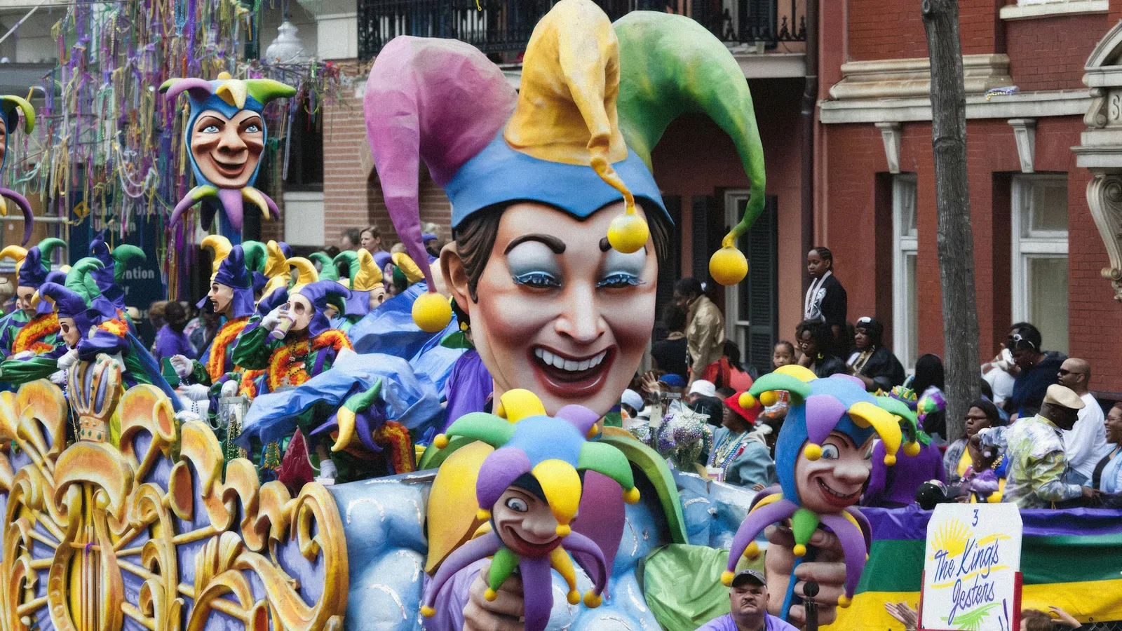 Colorful Mardi Gras parade float with large jester figures and festive crowd in the background on a city street.