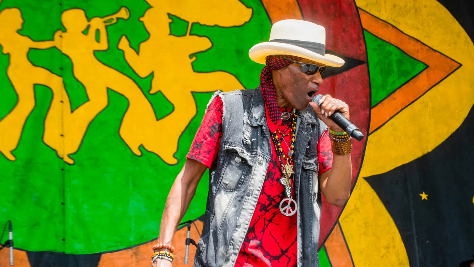 Male reggae singer wearing hat and sunglasses performs passionately on stage with colorful backdrop and band.