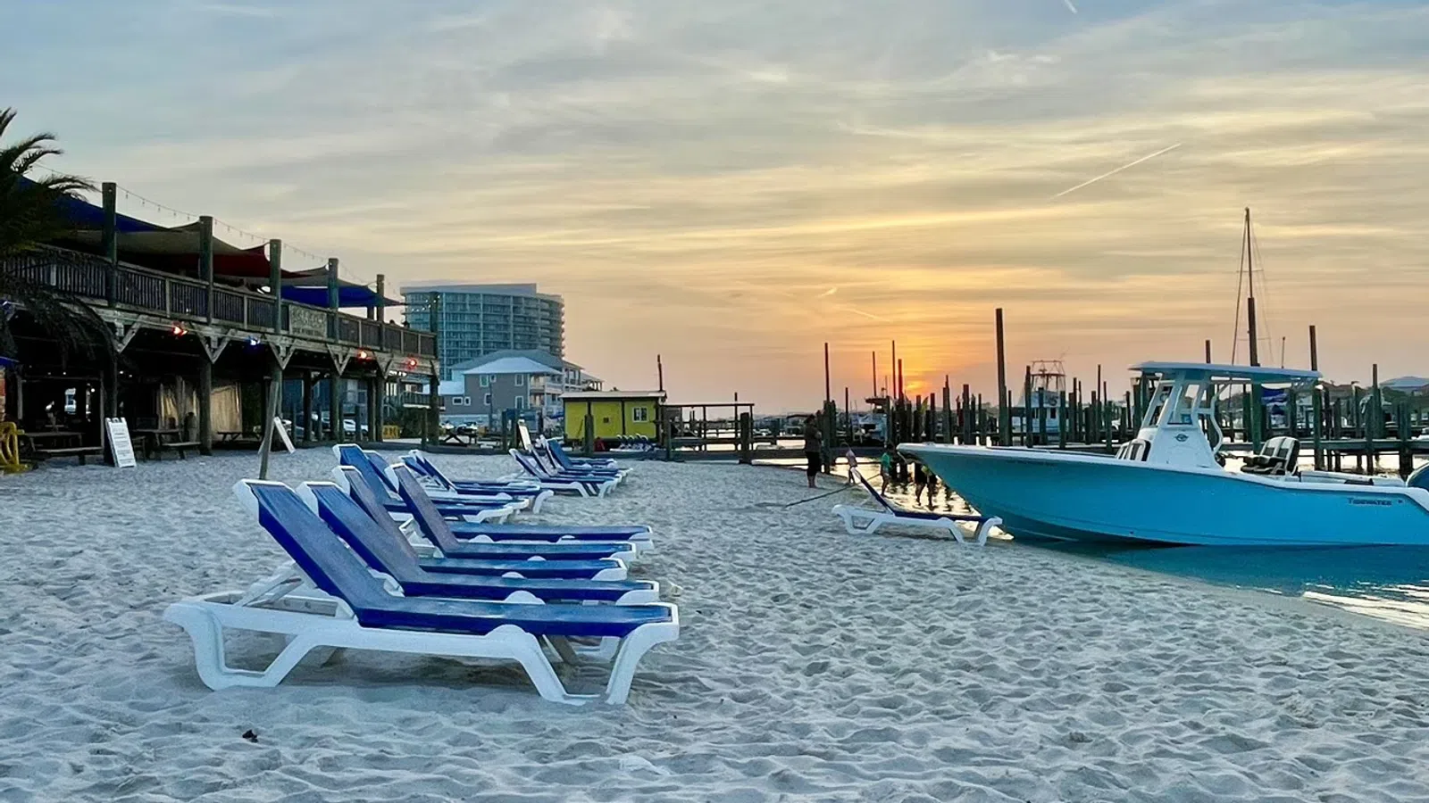 Empty blue and white beach chairs on sandy shore near docked boat at sunset with calm water and buildings.