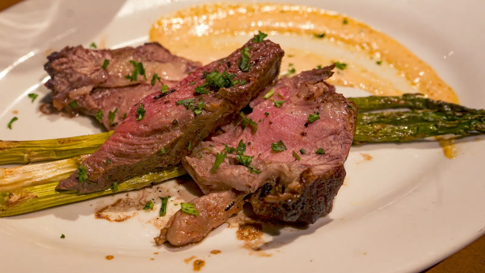 Grilled medium-rare steak slices served with asparagus and a creamy sauce on a white plate with herbs.