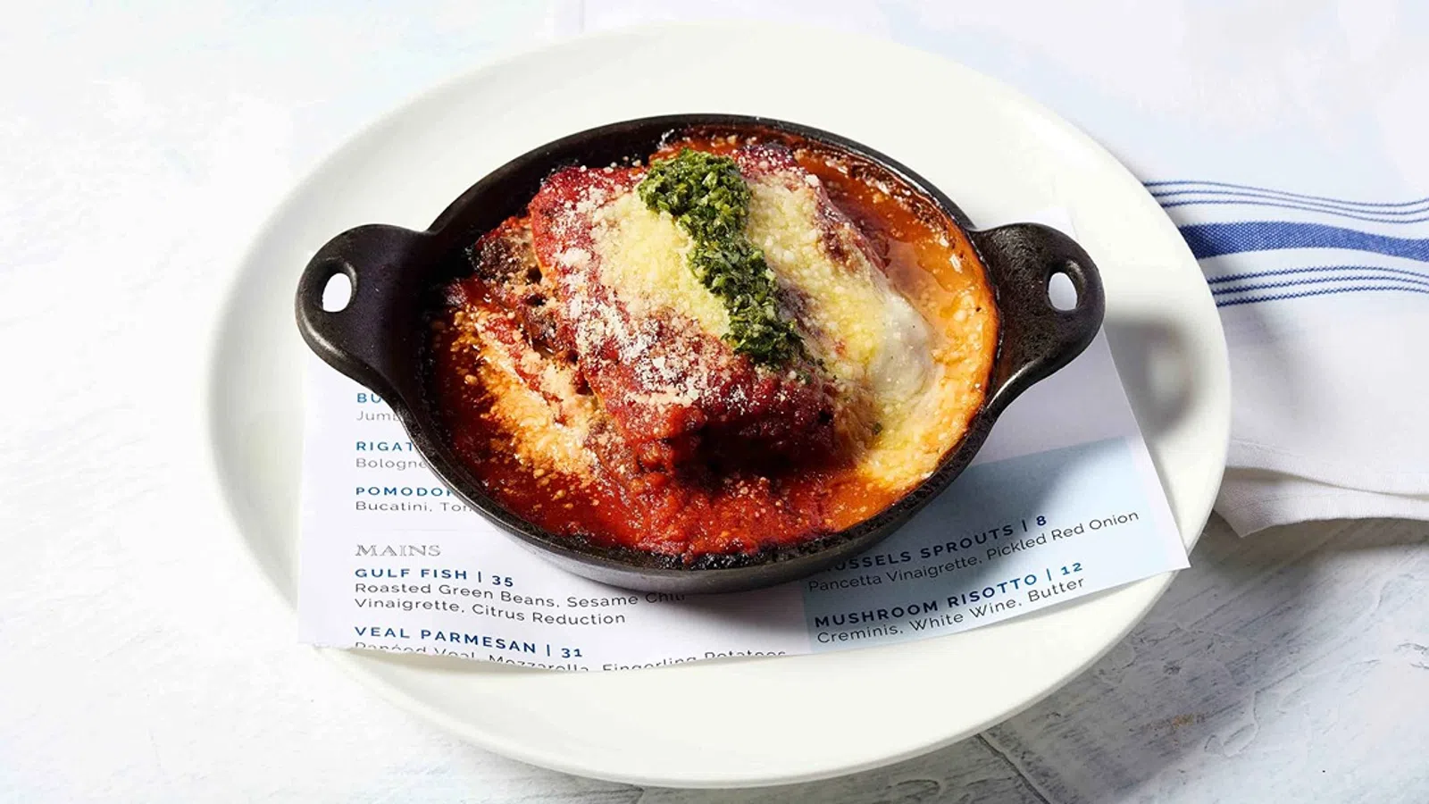 Baked veal parmesan with tomato sauce, cheese, and pesto served in a black dish on a white plate