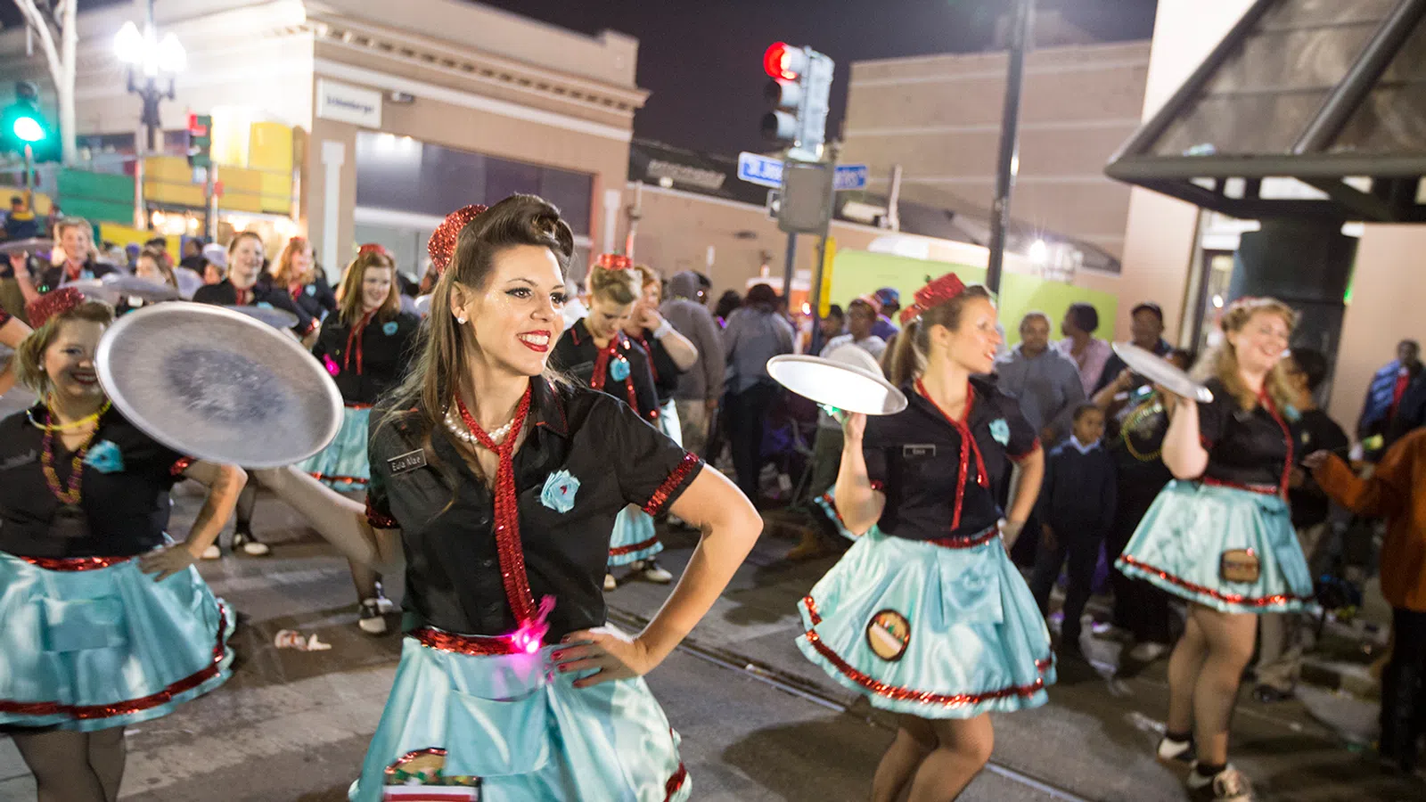 Women in vintage diner waitress costumes smiling and dancing with trays during a lively street parade at night.