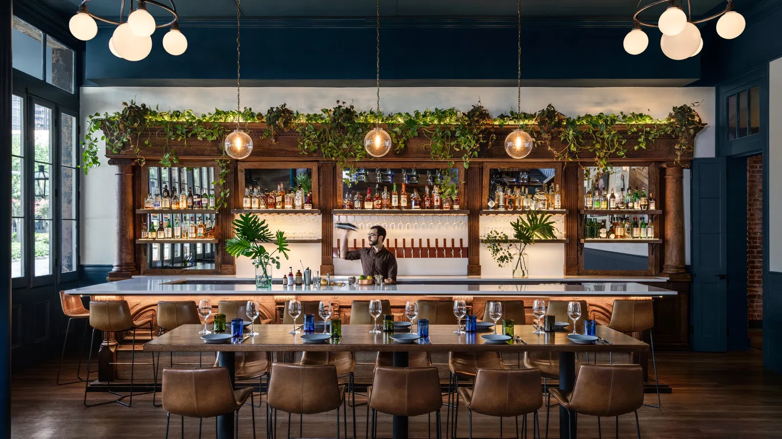 Stylish bar interior with wooden furniture, green plants, warm lighting, and a bartender preparing drinks behind the counter.