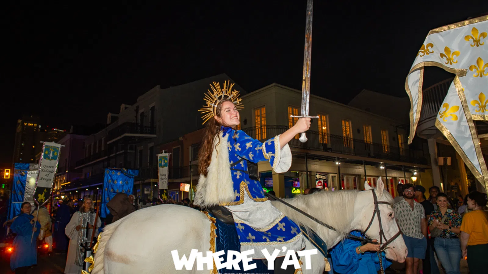 Person in royal costume holding sword on white horse during nighttime street parade with onlookers.