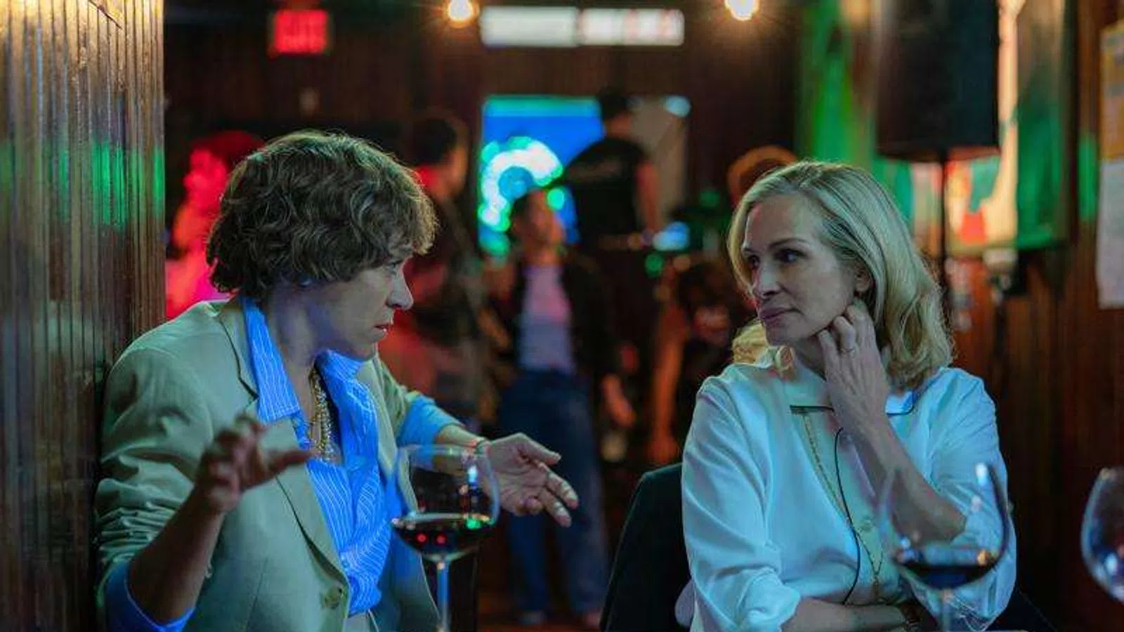 Two people engaged in conversation over wine glasses at a dimly lit bar with blurred background patrons.