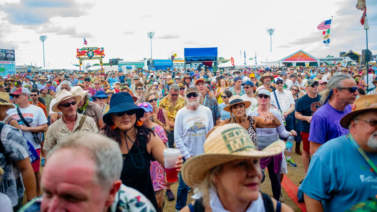 Large diverse crowd at an outdoor festival under a cloudy sky with colorful tents and flags.