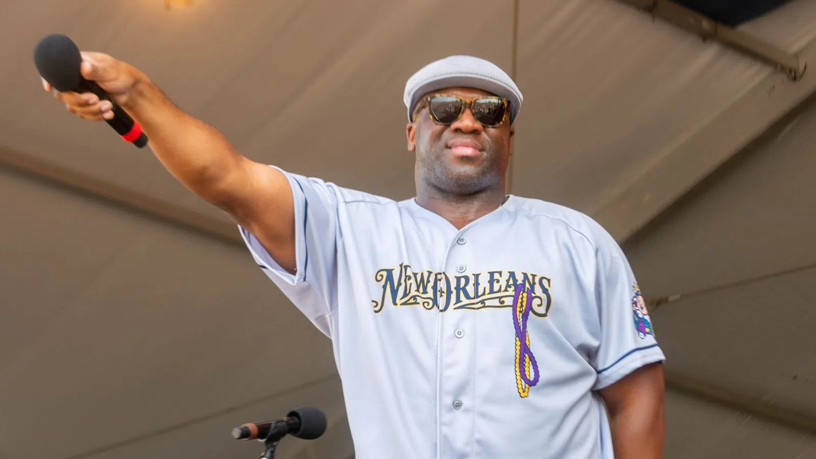Man in New Orleans jersey and sunglasses holds microphone on stage under tent ceiling