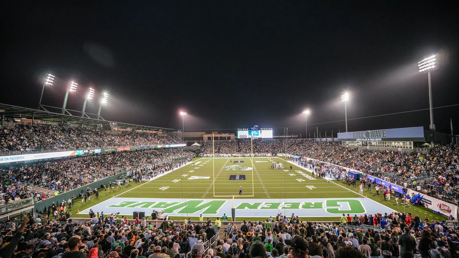 Nighttime football game with full stadium and bright floodlights illuminating the field and crowd.