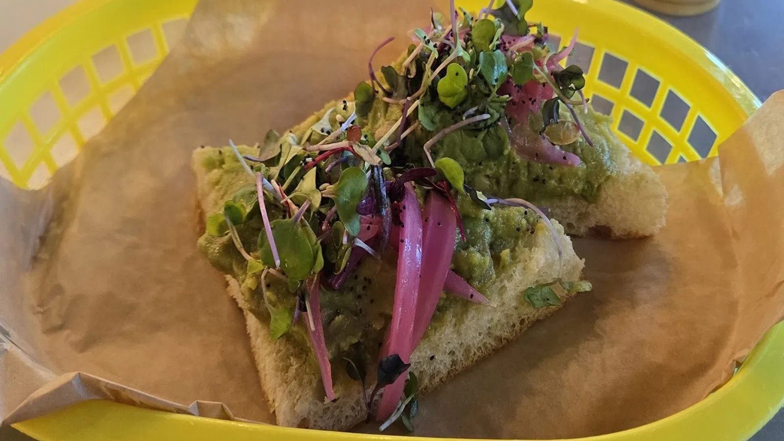 Open-faced sandwich with guacamole, pickled red onions, and microgreens on focaccia bread in a yellow basket.