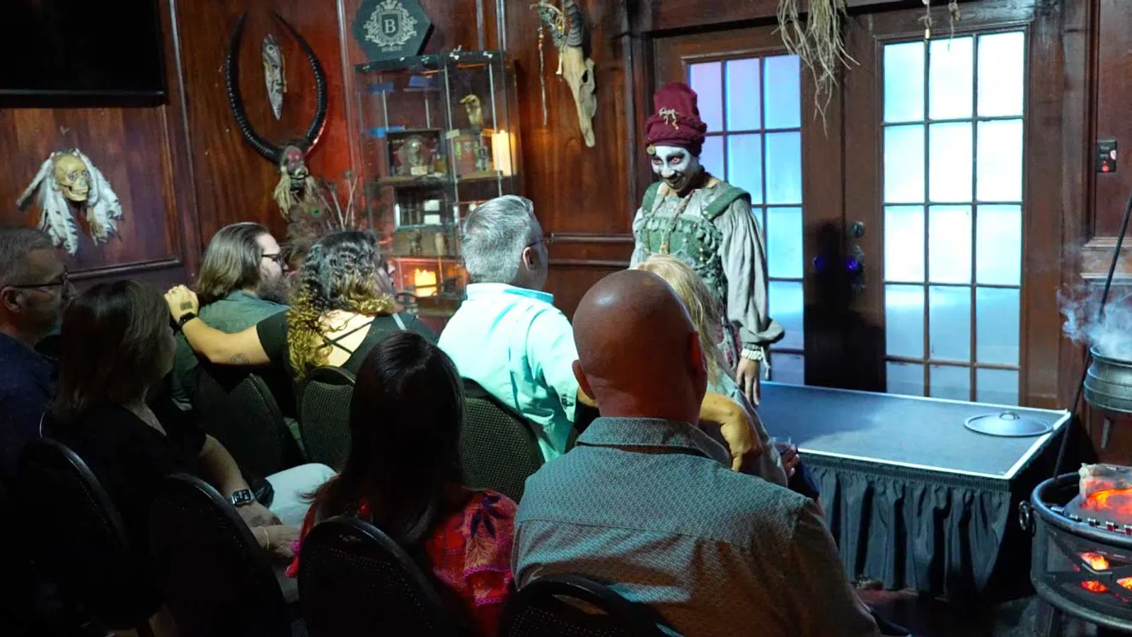 Audience watches performer in creepy costume and makeup on stage in dimly lit room with spooky decorations.