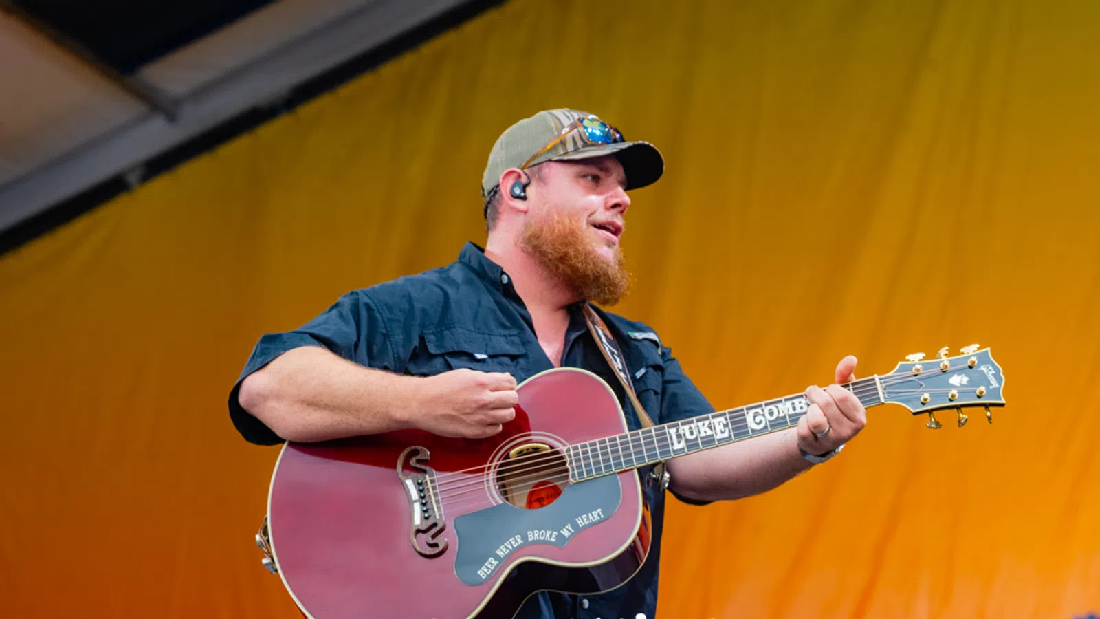 Male country musician playing guitar on stage with a yellow backdrop and wearing a cap and sunglasses