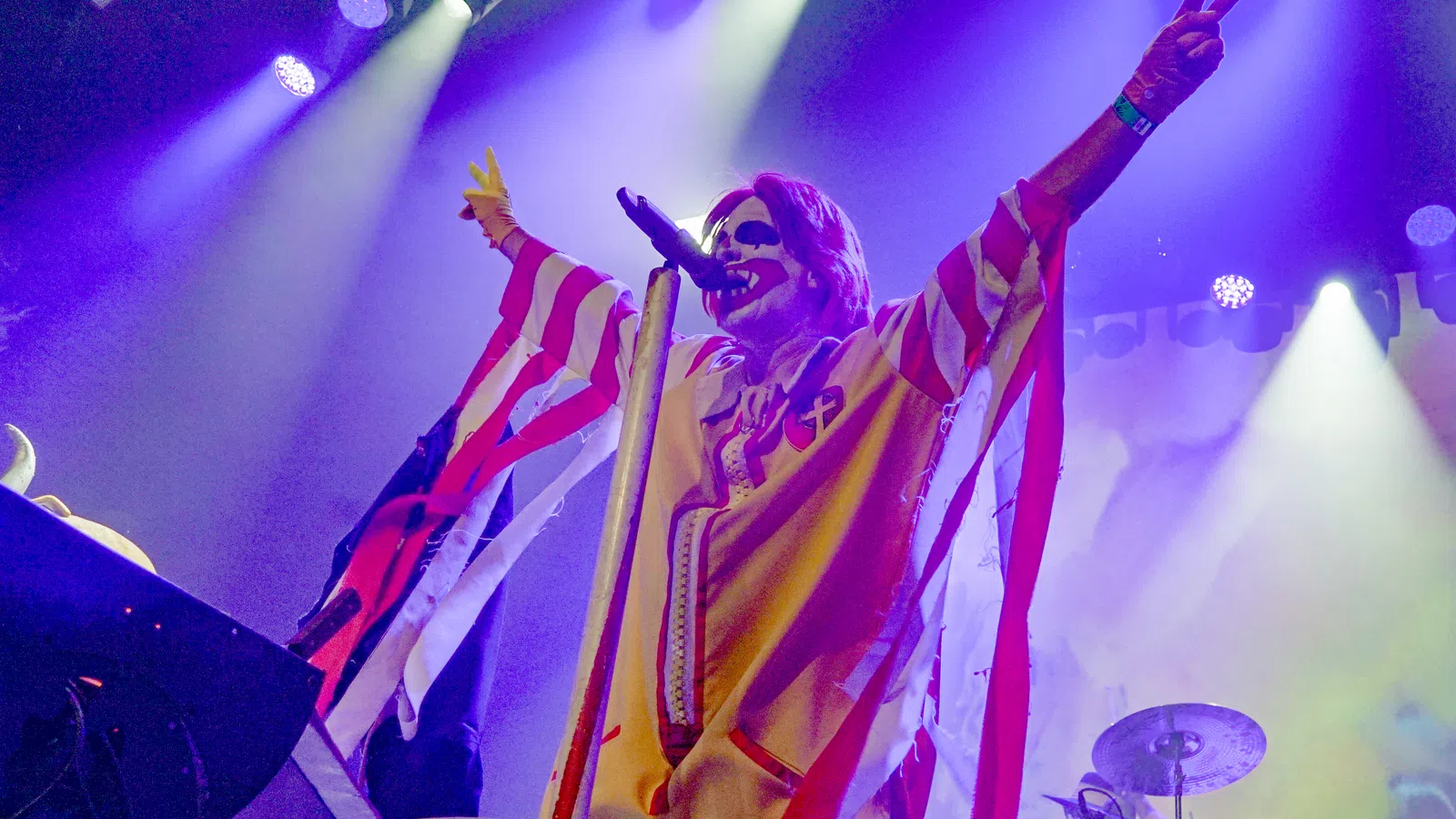 A performer in a vivid clown costume strikes a pose on stage with colorful lighting.