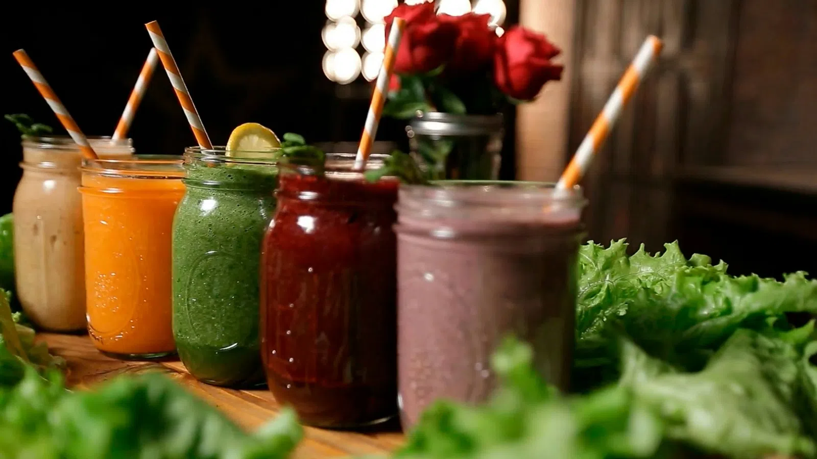 Colorful smoothies in glass jars with striped straws lined up on wood board with fresh greens and red roses