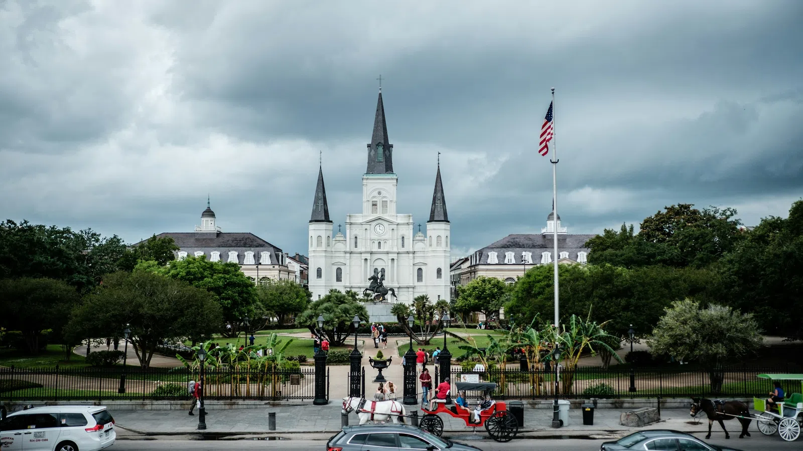 St. Louis Cathedral in New Orleans with horse carriages, cars, American flag, and cloudy sky in the background