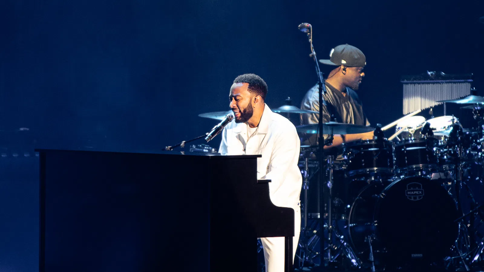 Singer playing piano and drummer performing on stage with dark blue background during concert