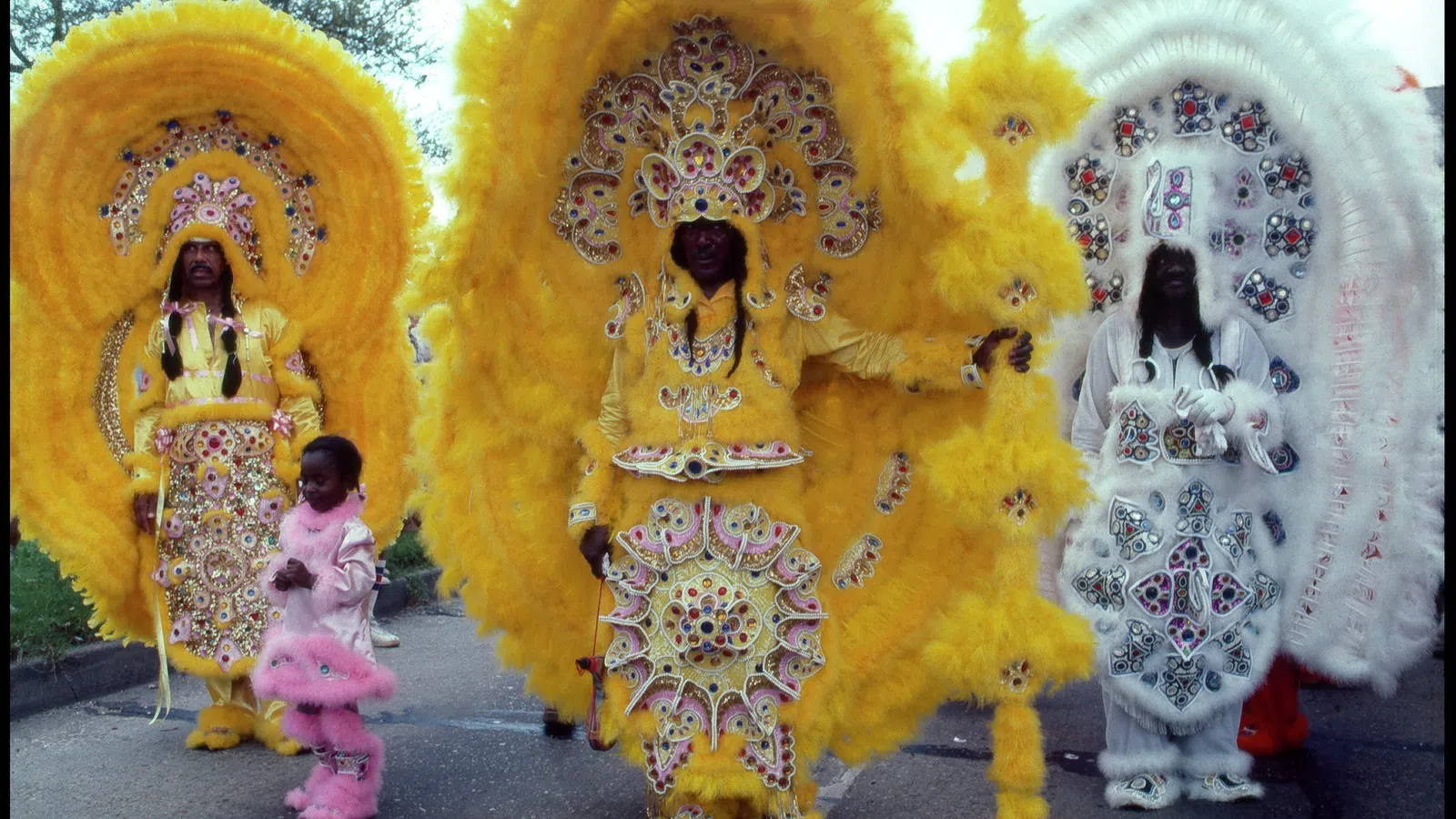 Three people in elaborate yellow and white beaded Mardi Gras costumes with large feathered headdresses stand outdoors