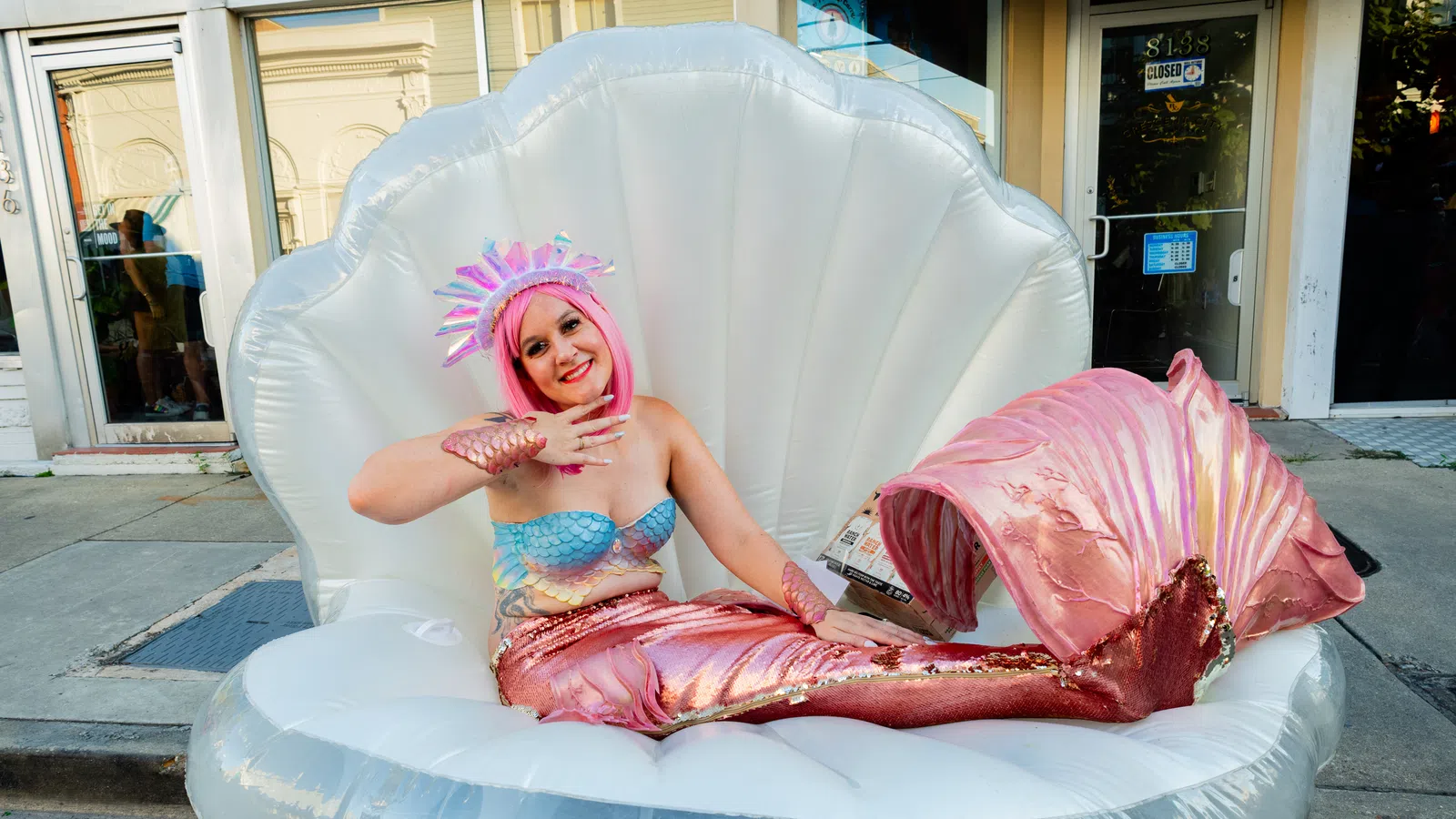 Smiling woman in a pink mermaid costume sitting on a large inflatable clam shell on a city street.