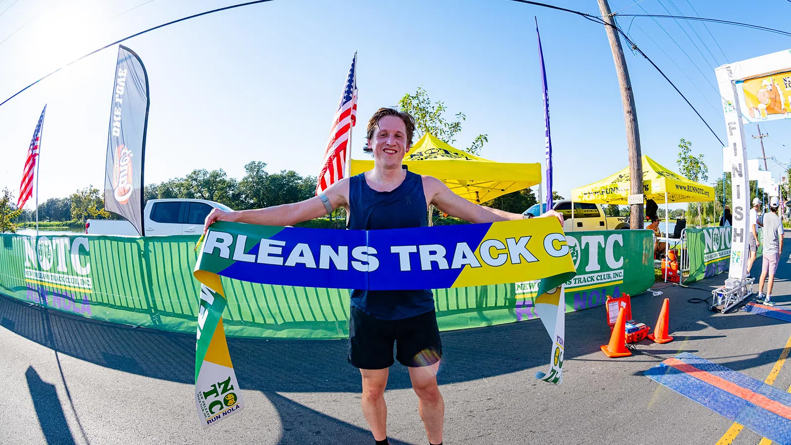 Runner celebrates victory holding Orleans Track Club banner at outdoor race finish line on sunny day