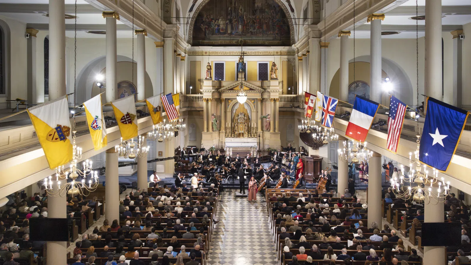 Interior of a historic church filled with an audience attending a musical concert with flags hanging along the walls.