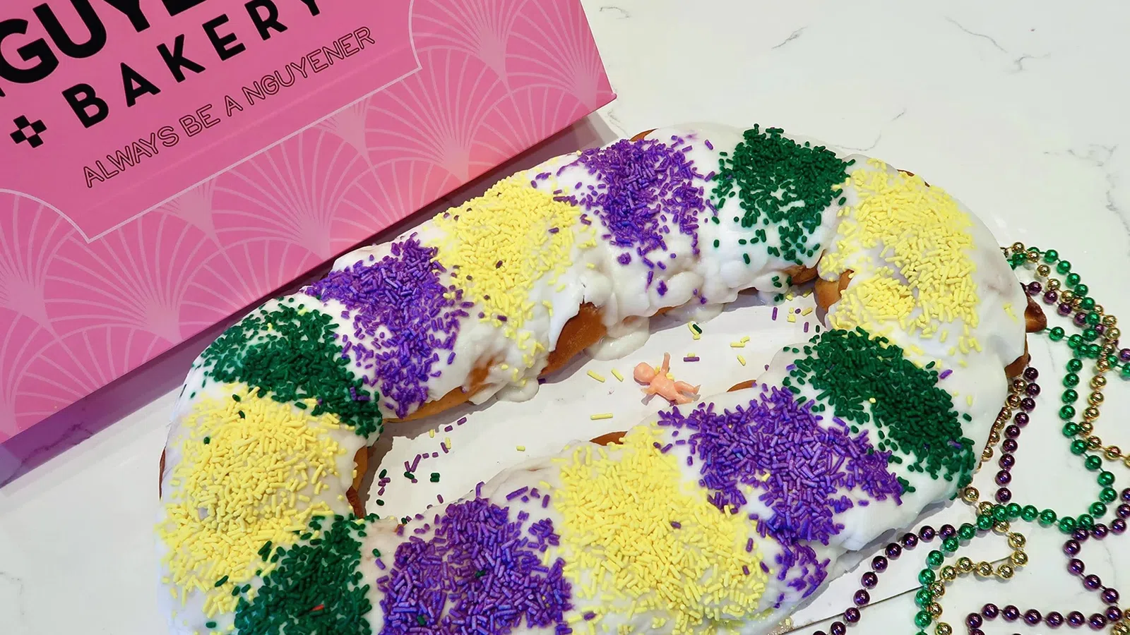 King cake with white icing and purple, yellow, green sprinkles next to pink Douceur Guyenere's Bakery box