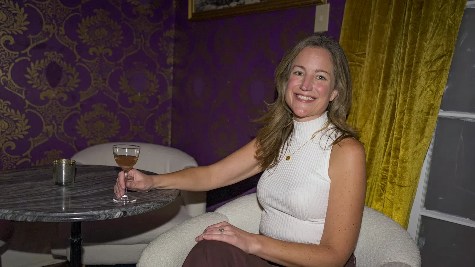 Smiling woman in white sleeveless top sitting in armchair holding a cocktail glass at a marble table