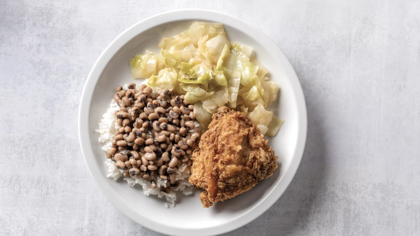 Plate with fried chicken, cooked cabbage, and black-eyed peas over white rice on a light background.