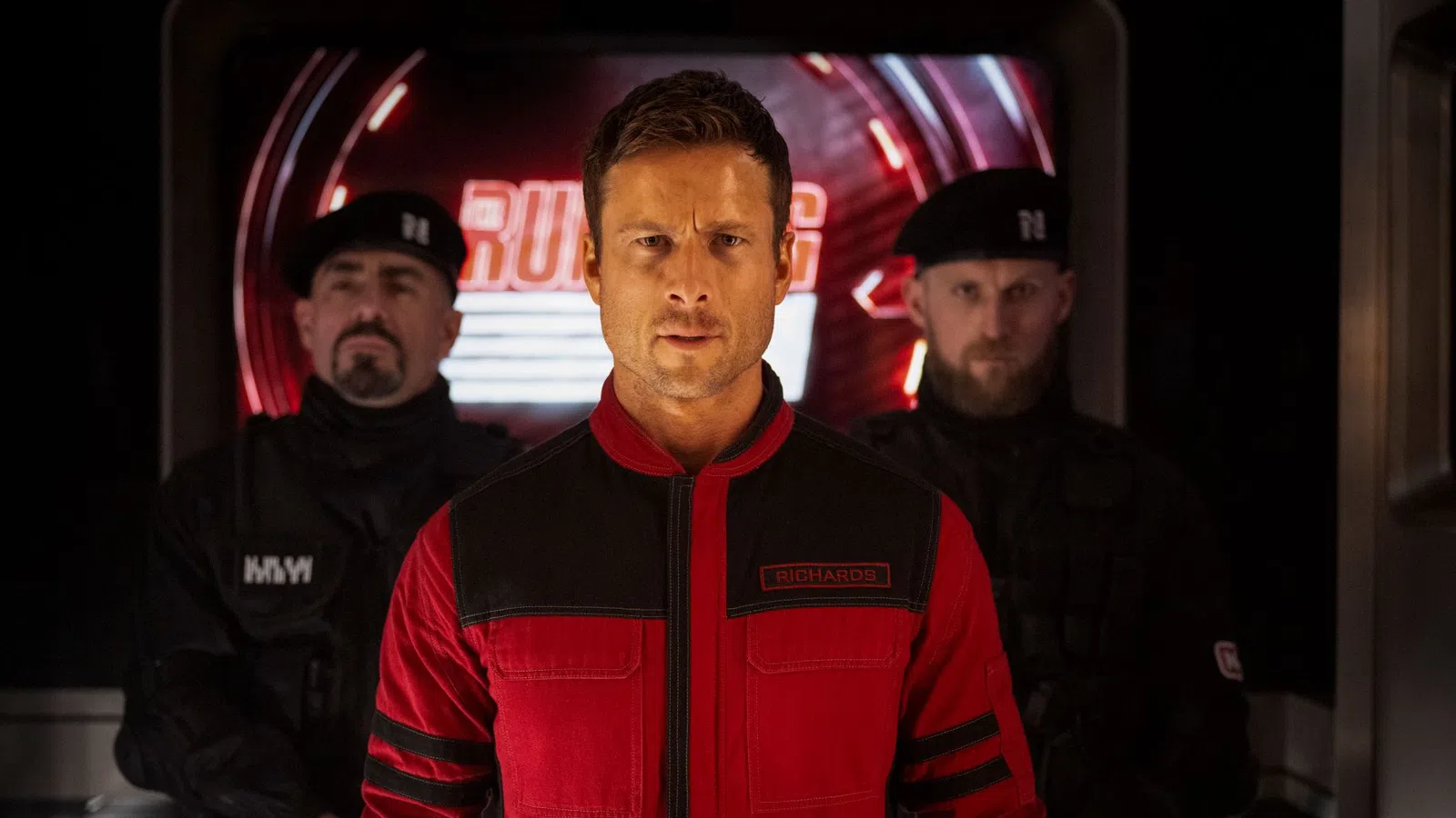 Man in red and black jacket with two guards in black uniforms standing behind him under dim lighting.