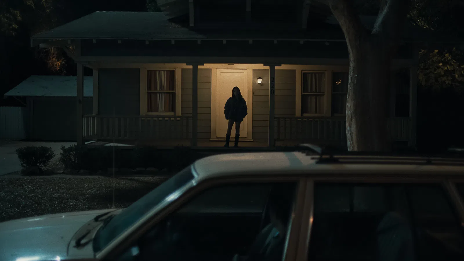 Person standing in the lit doorway of a house at night with a parked car in the foreground.